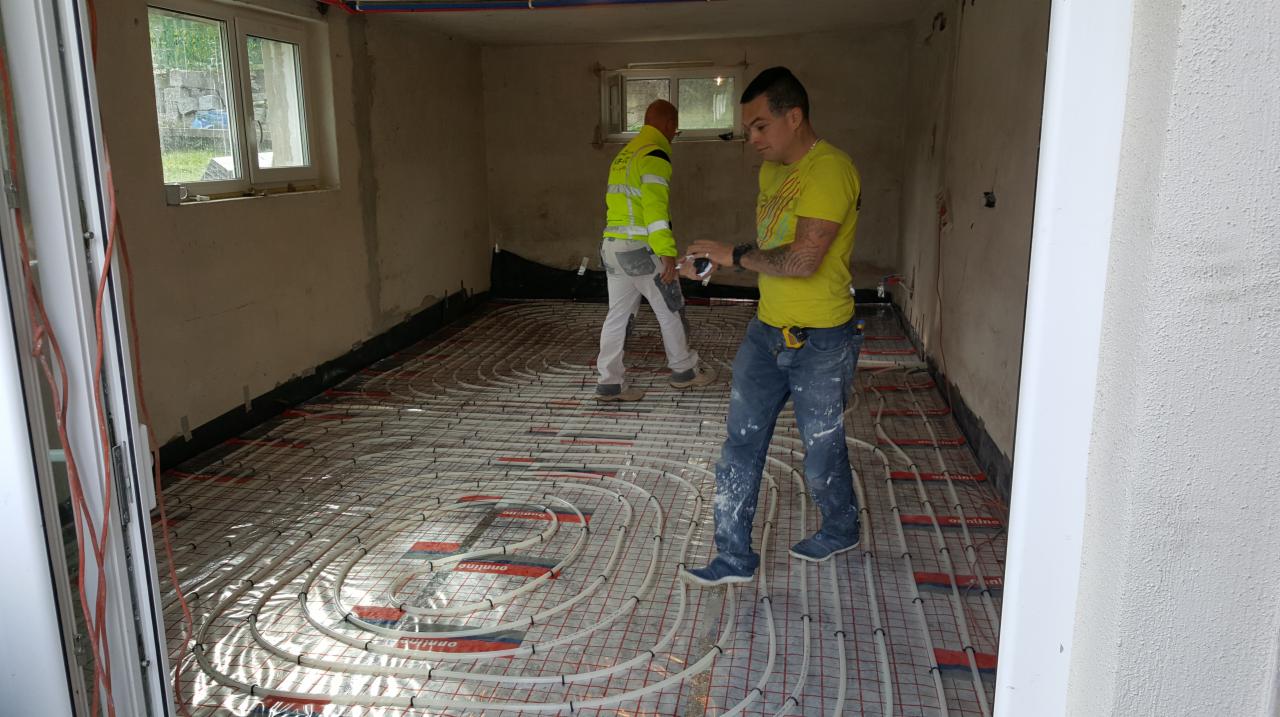 Dwóch instalatorów montuje ogrzewanie podłogowe w surowym pomieszczeniu z widocznym oknem i elementami instalacji na ścianach.