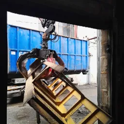 Chwytak hydrauliczny podnosi metalowy element konstrukcyjny na tle niebieskiego kontenera, widok z wnętrza hali.