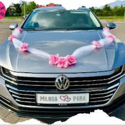 Srebrny samochód udekorowany białym tiulem i różowymi kwiatami na masce, tablica rejestracyjna z napisem 'Młoda Para' i dwoma czerwonymi sercami, widok z przodu na tle ulicy.