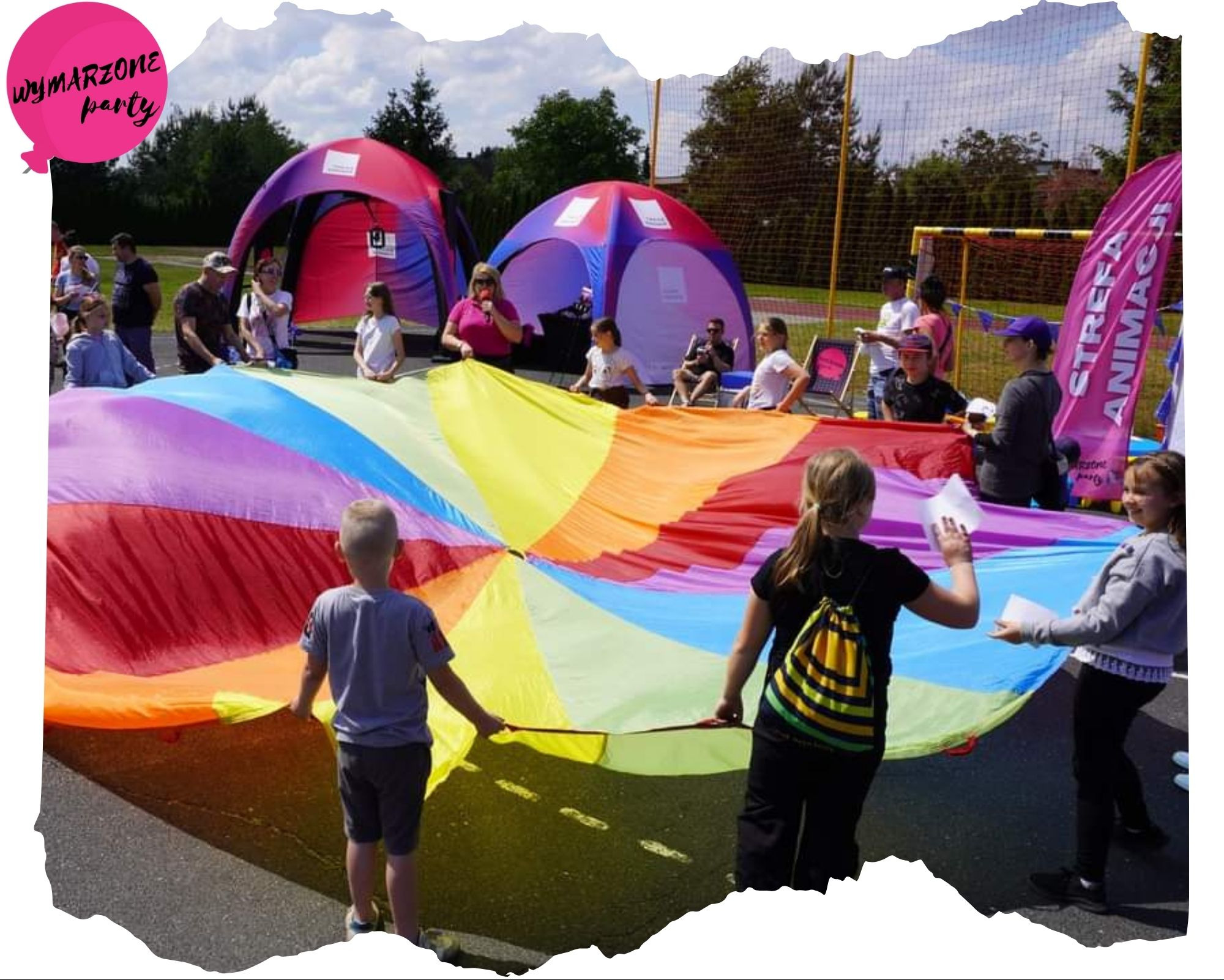 Dzieci trzymają wielobarwną chustę animacyjną podczas zabawy na świeżym powietrzu, w tle namioty i osoby dorosłe.