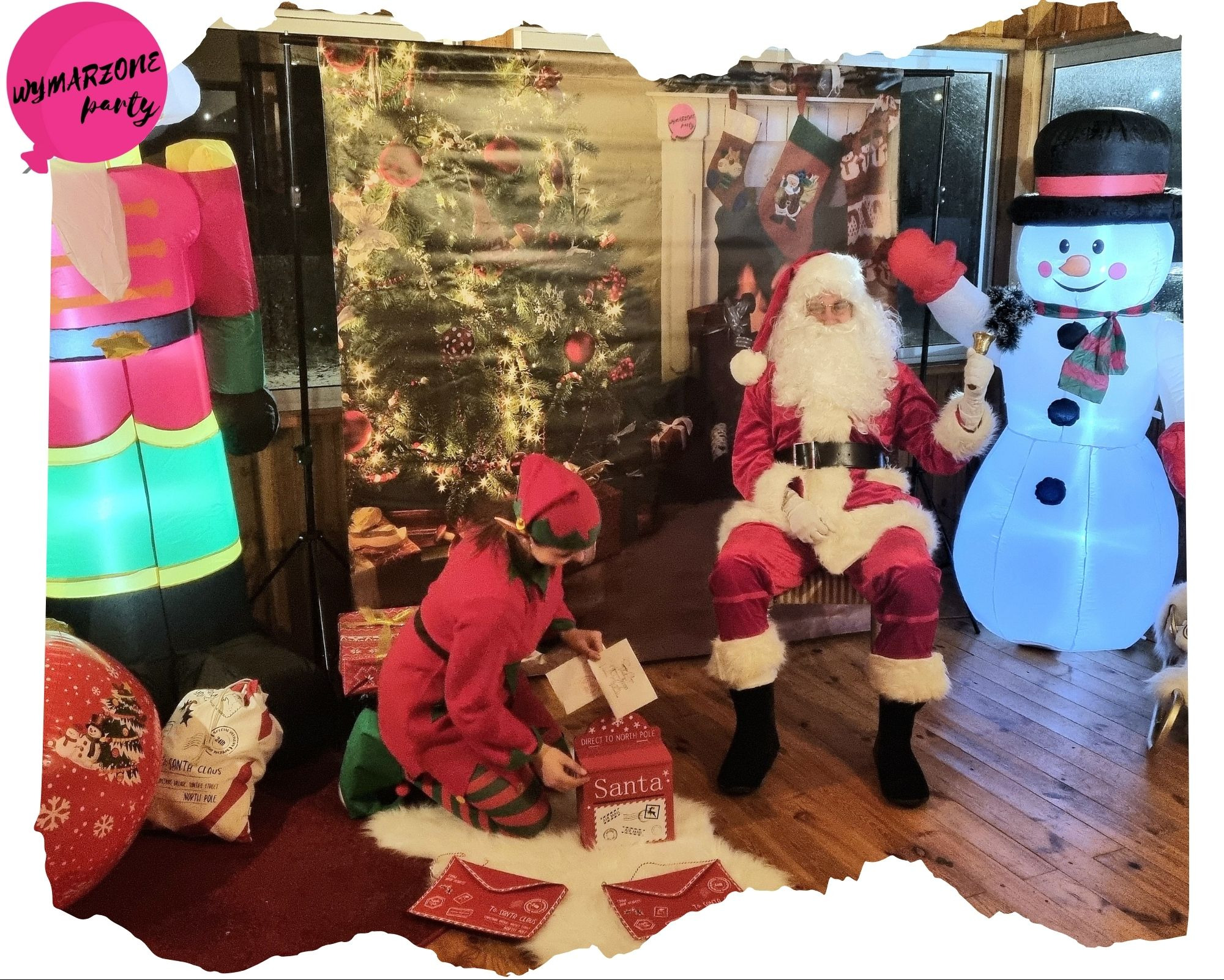 Inscenizacja świąteczna: Mikołaj siedzi obok dmuchanego bałwana, elf czyta listy z czerwonej skrzynki 'Santa', w tle choinka i dmuchany dziadek do orzechów.