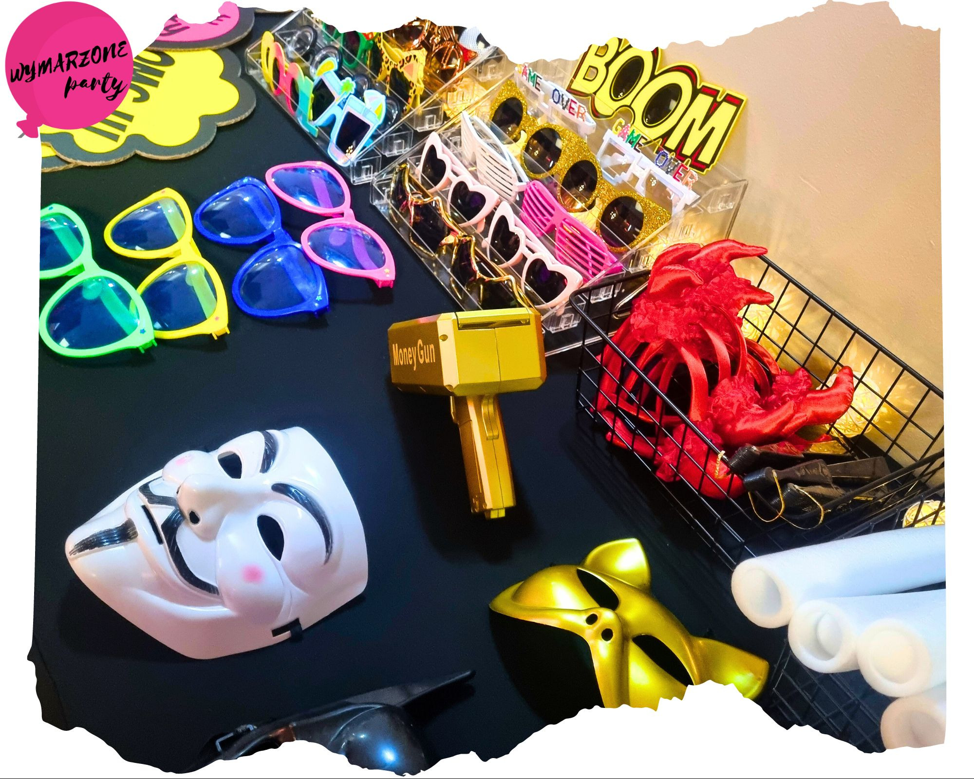 Różnorodność rekwizytów imprezowych na czarnym tle: okulary przeciwsłoneczne w różnych kolorach i kształtach, maski anonima i złoty hełm, złoty pistolet na pieniądze, czerwone ręce szkieletu...