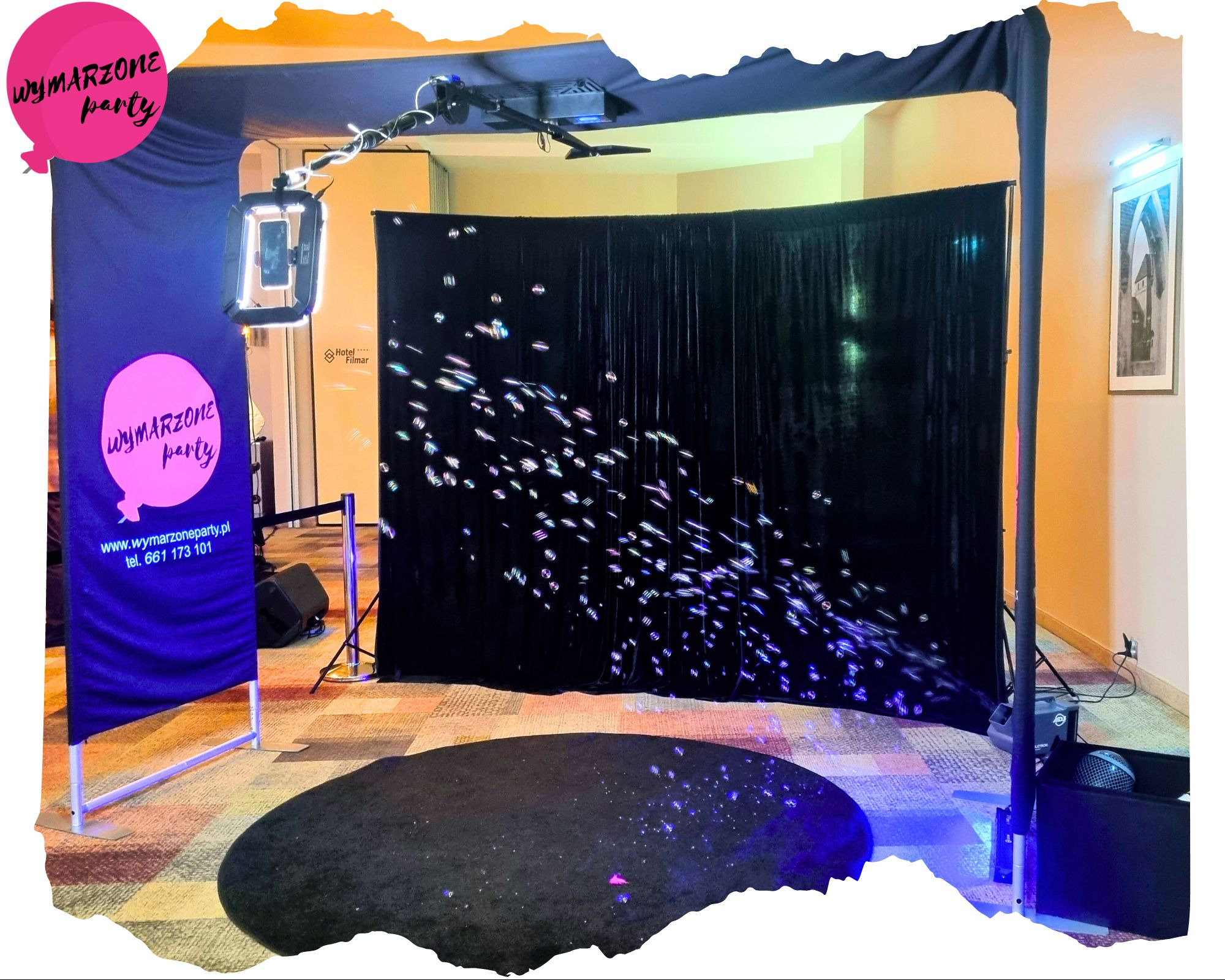 Rozstawiona fotobudka z czarnym tłem i efektami świetlnymi w pomieszczeniu, obok baner reklamowy z logo firmy.