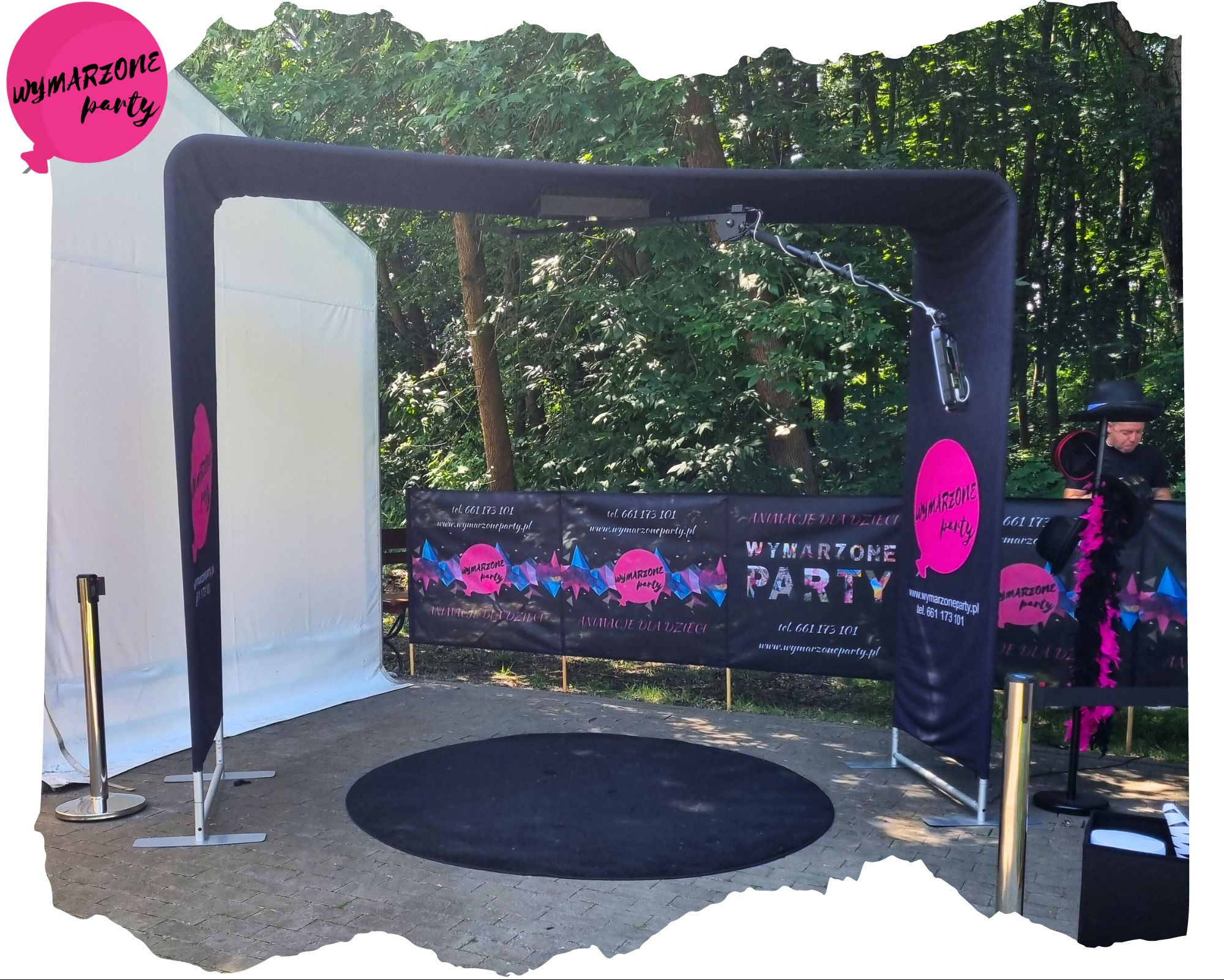 Rozstawiona fotobudka z czarnym tłem w kształcie koła, na zewnątrz, z widocznym operatorem i banerem reklamowym firmy 'Wymarzone Party' w tle.