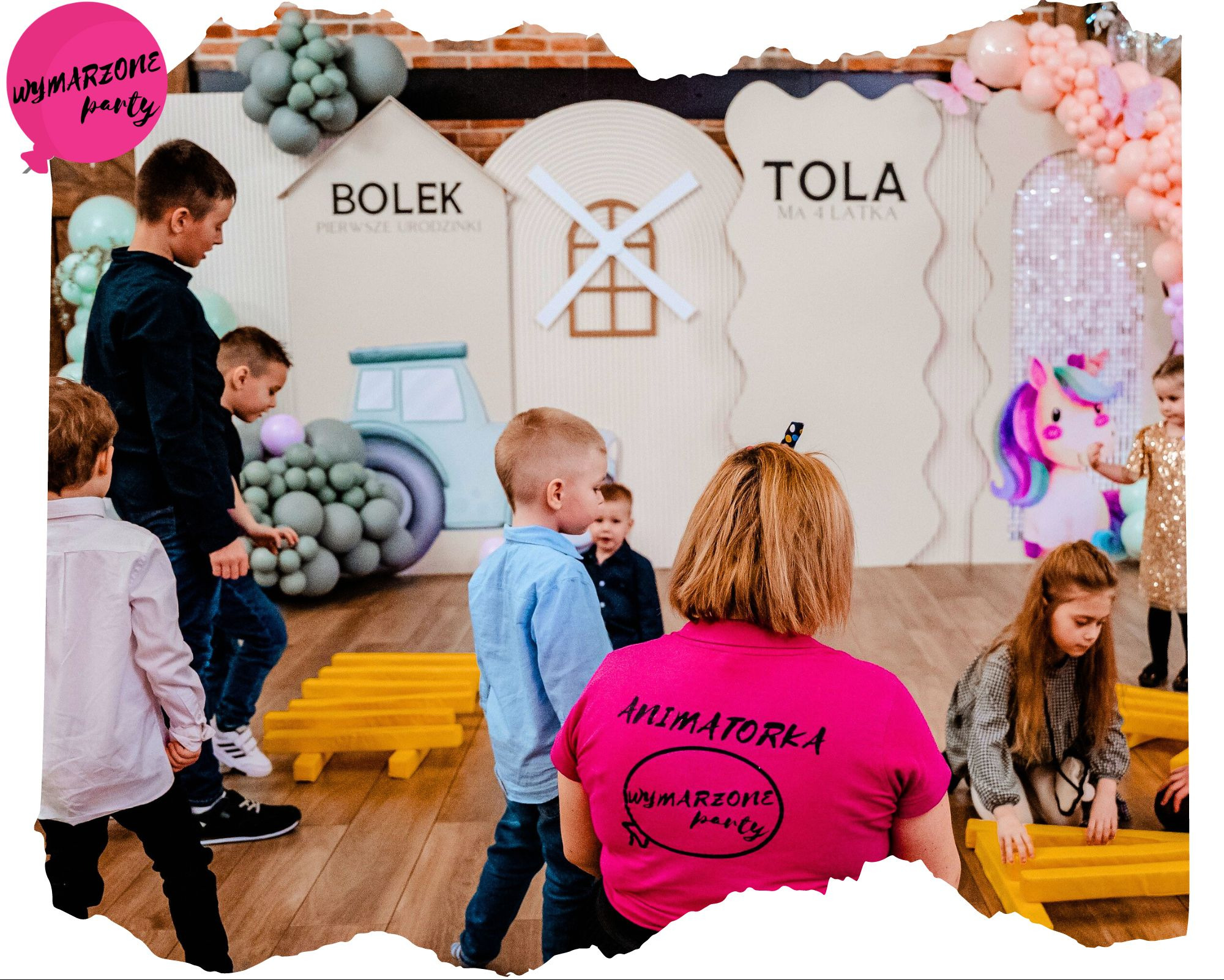 Urodzinowa dekoracja z motywem traktora i wiatraka z balonów oraz dzieci bawiące się pod okiem animatorki w różowej koszulce z napisem.