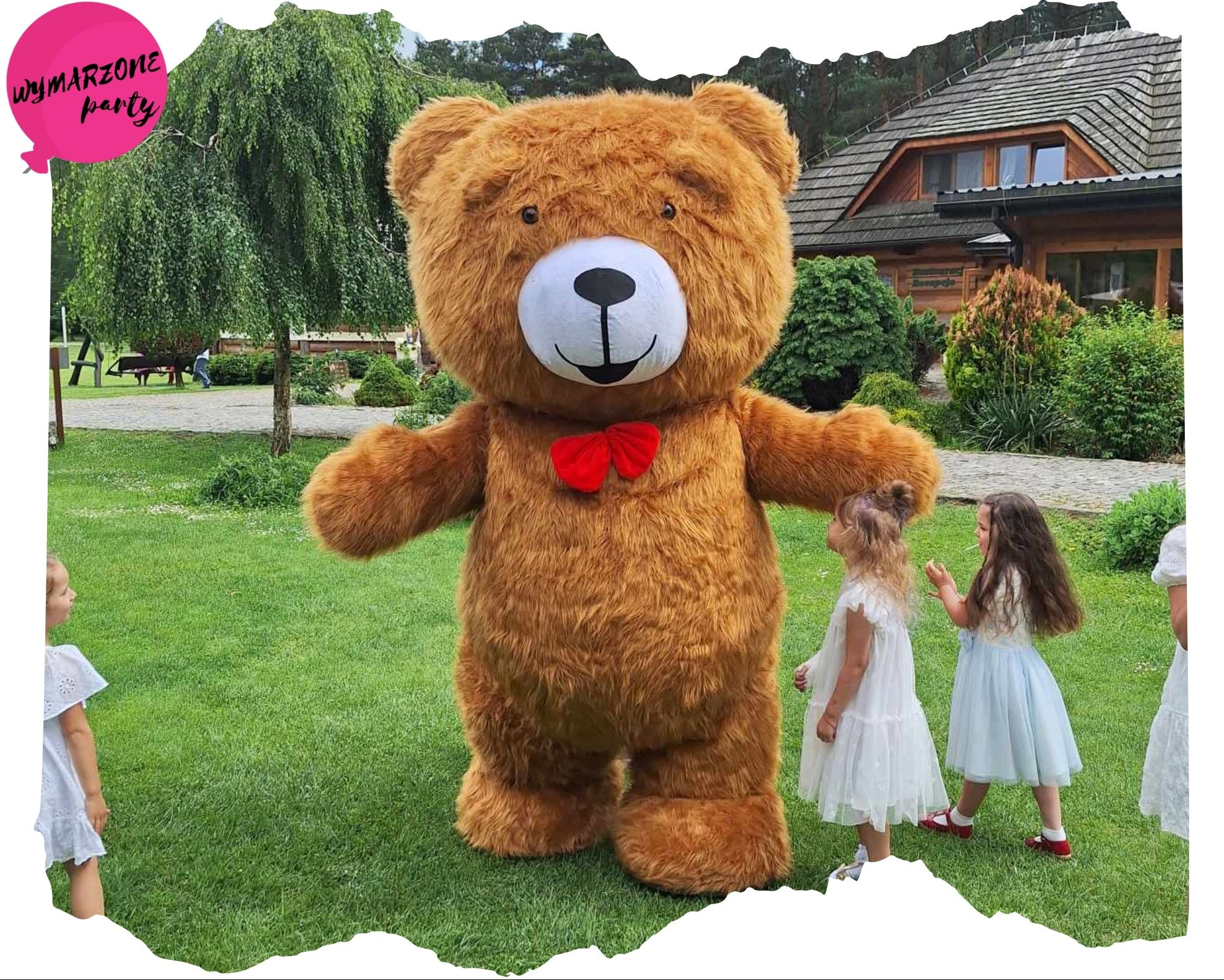 Duży, pluszowy miś z czerwoną muchą, otoczony przez grupę dziewczynek w białych sukienkach na zielonym trawniku przed drewnianym budynkiem.