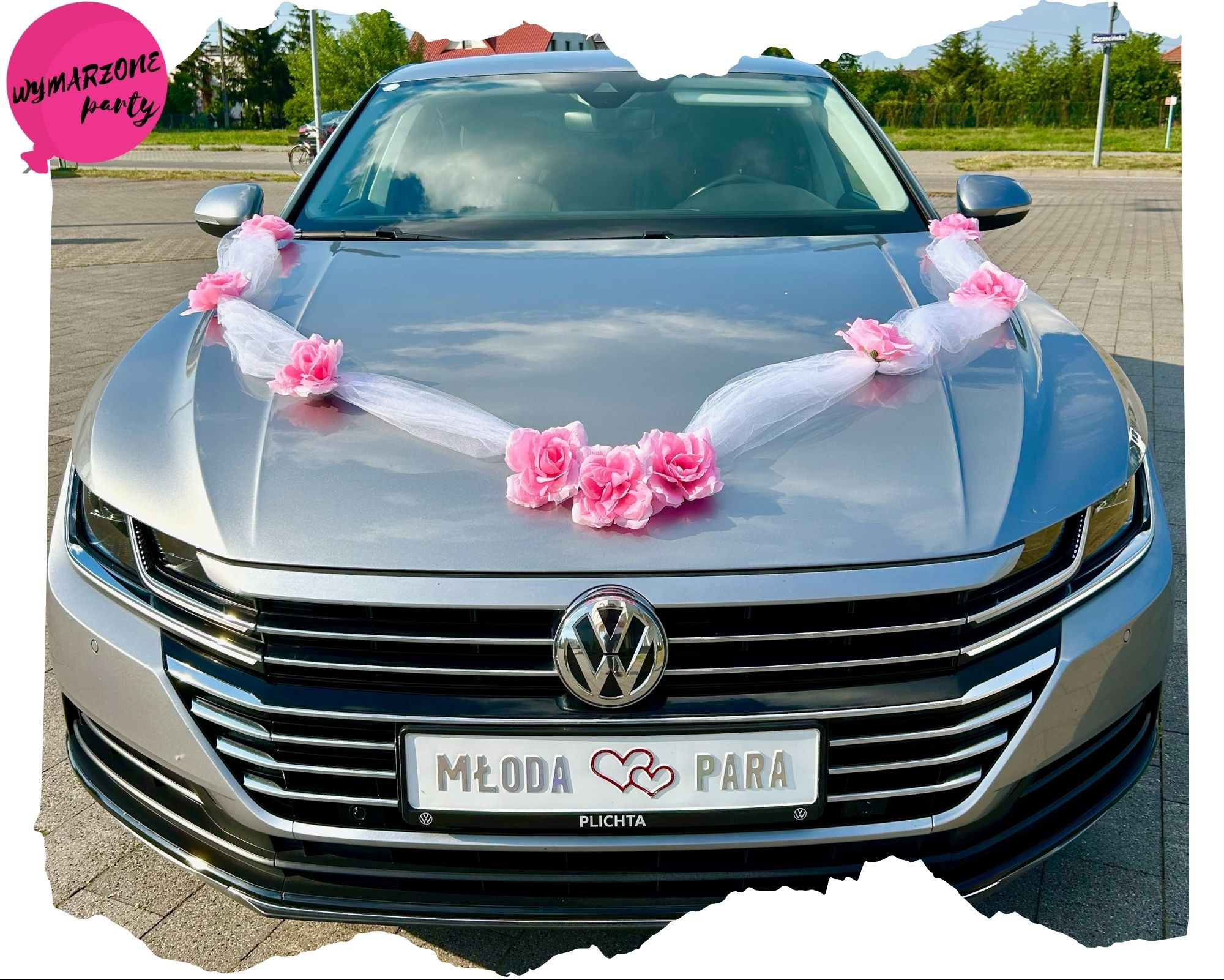 Srebrny samochód udekorowany białym tiulem i różowymi kwiatami na masce, tablica rejestracyjna z napisem 'Młoda Para' i dwoma czerwonymi sercami, widok z przodu na tle ulicy.