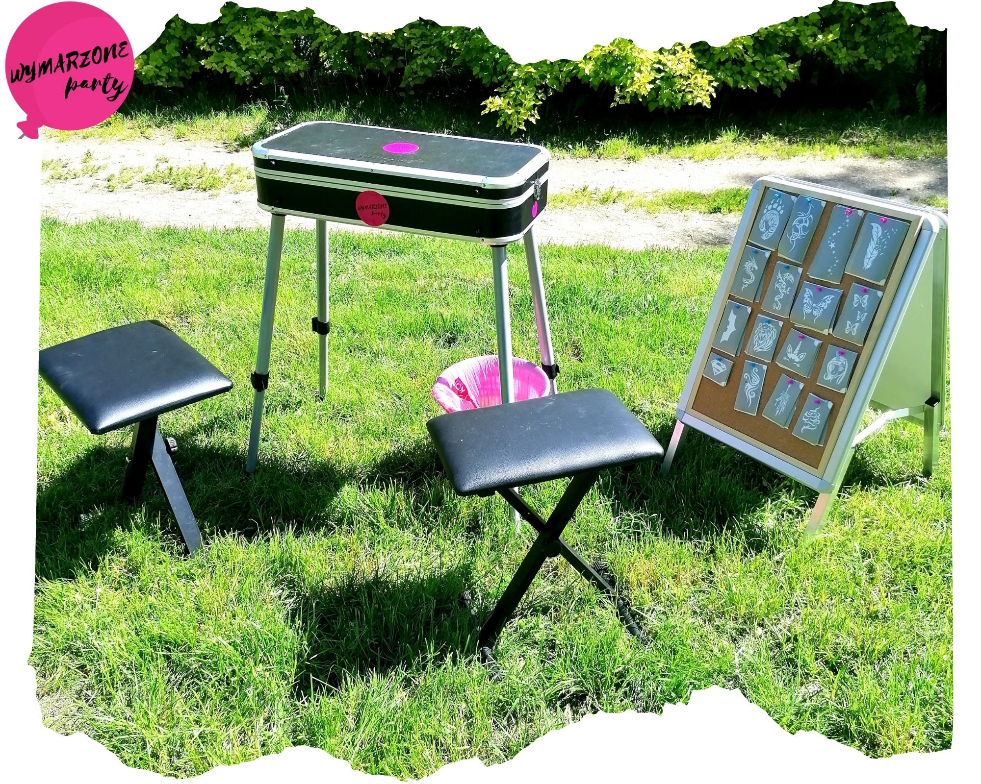 Stanowisko do wykonywania tatuaży brokatowych na trawie: stolik z logo firmy, tablica z wzorami tatuaży i dwa stołki.
