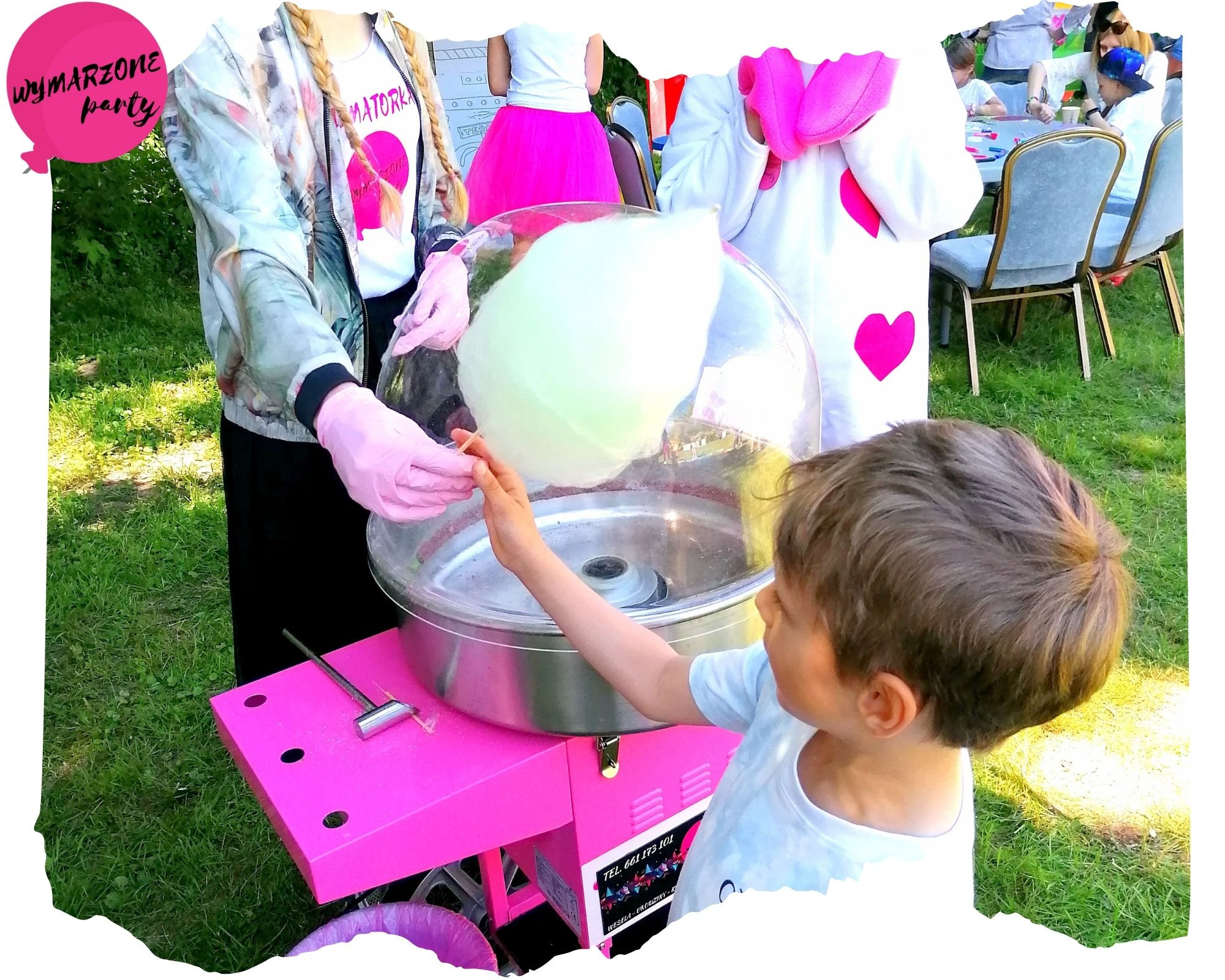 Chłopiec wyciąga rękę po watę cukrową, którą podaje osoba w różowych rękawiczkach przy różowej maszynie do waty cukrowej na trawie, w tle dzieci przy stole i osoba w kostiumie.