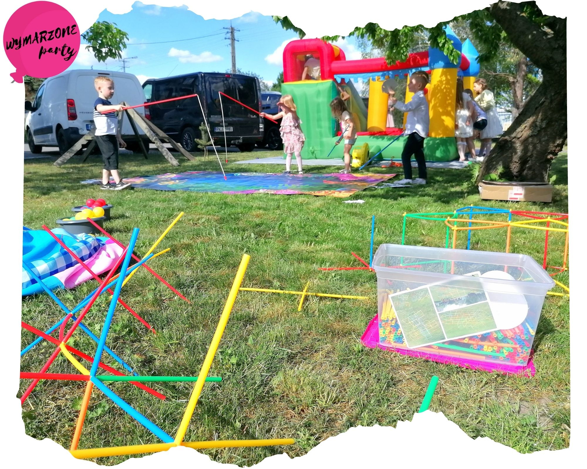 Dzieci bawiące się na trawie podczas imprezy plenerowej z dmuchanym zamkiem w tle, wykorzystując kolorowe rurki do budowy konstrukcji oraz matę do malowania wodą.