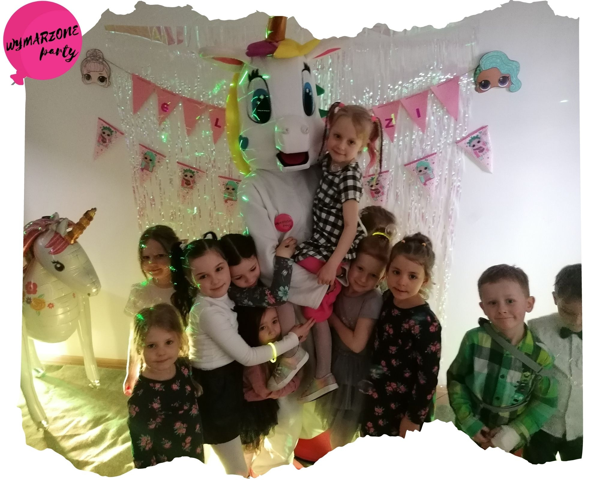 Uśmiechnięte dzieci otaczają osobę w kostiumie jednorożca z podświetlanymi elementami, balon w kształcie jednorożca i dekoracje urodzinowe w tle.