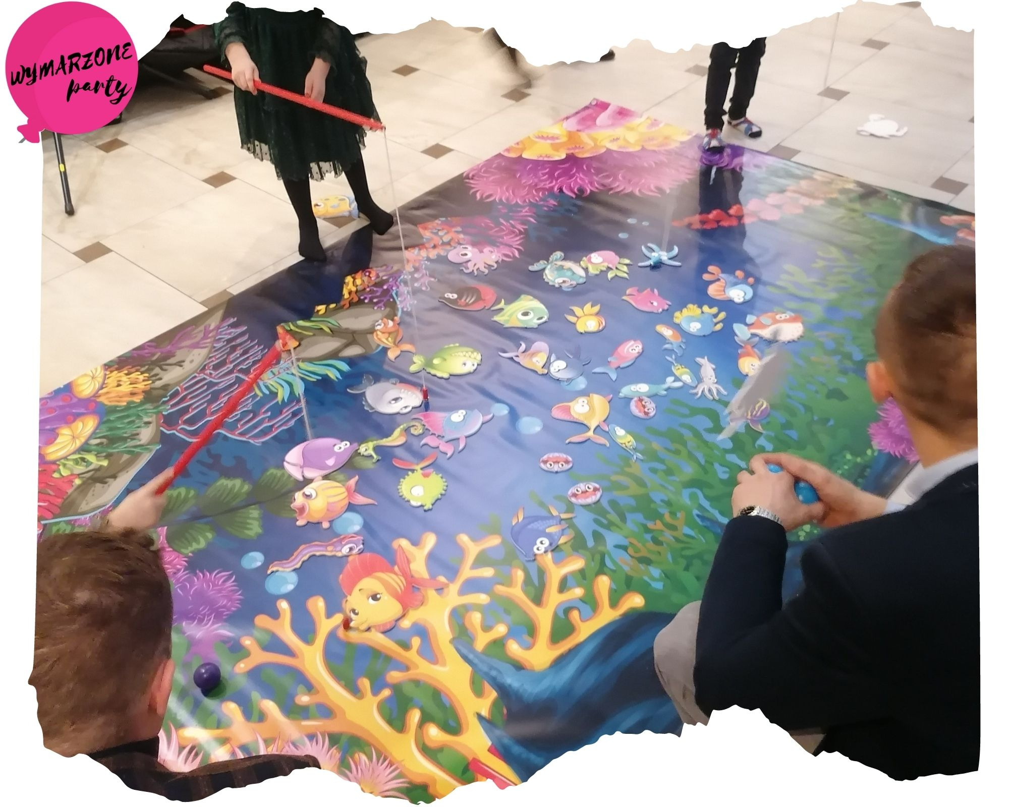 Dzieci w Toruniu bawią się na dużej, kolorowej macie z motywem podwodnego świata, łowiąc rybki za pomocą wędek i plastikowych narzędzi.