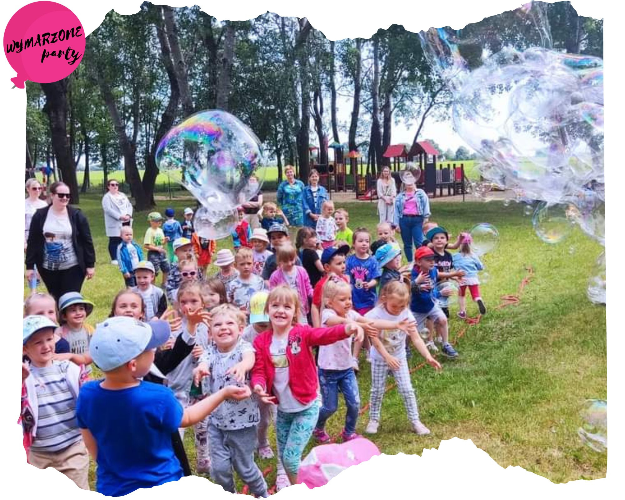 Grupa roześmianych dzieci na trawie bawi się w puszczanie ogromnych baniek mydlanych na tle placu zabaw i drzew.