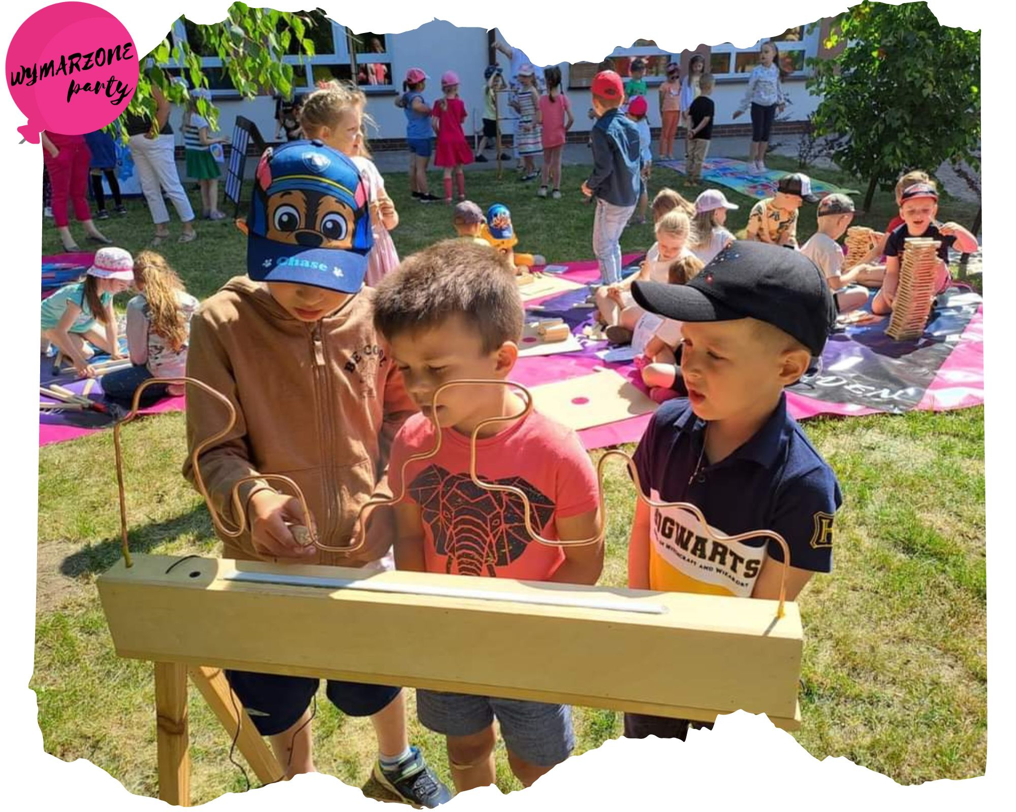 Trójka dzieci skupiona na grze zręcznościowej z metalowym labiryntem na drewnianym stelażu, w tle grupa dzieci na imprezie plenerowej.