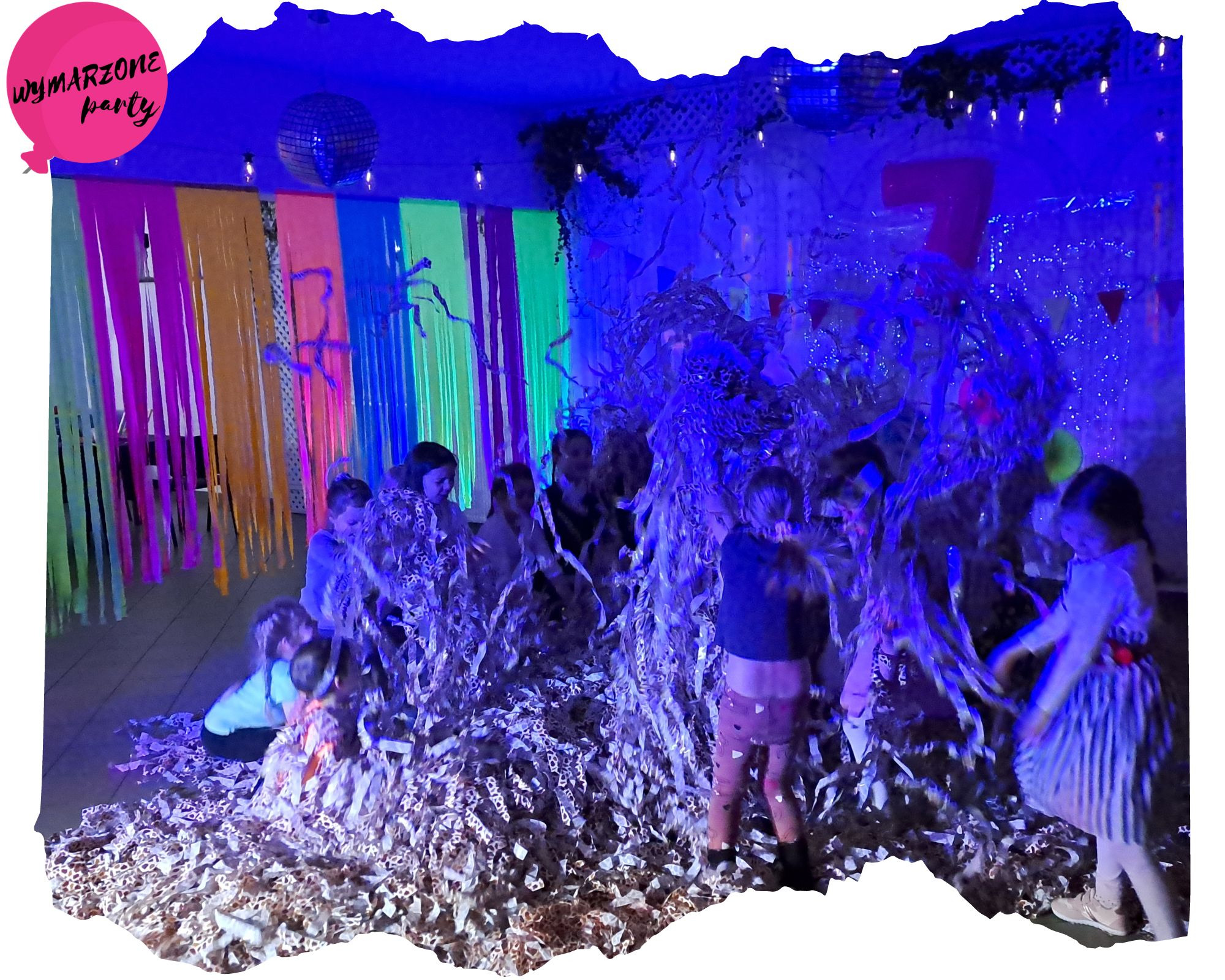 Grupa dzieci bawi się w stercie konfetti w pomieszczeniu udekorowanym kolorowymi wstążkami i dyskotekową kulą, podświetlonym niebieskim światłem.