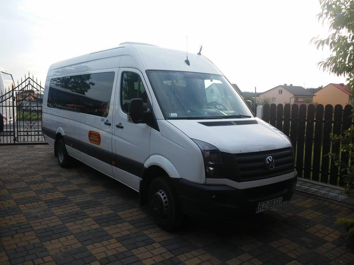 Biały bus Volkswagen Crafter z przyciemnionymi szybami, zaparkowany na kostce brukowej przed czarnym metalowym ogrodzeniem i zielenią.