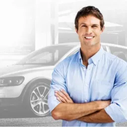 Uśmiechnięty mężczyzna w błękitnej koszuli z założonymi rękami, na rozmytym tle samochodu i miejskiej ulicy, obok logo firmy ubezpieczeniowej.