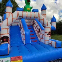 Nadmuchiwany zamek ze zjeżdżalnią, ozdobiony motywami z bajki Moana i figurą zielonego dinozaura na szczycie, na tle błękitnego nieba z chmurami.