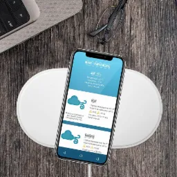 Smartfon z uruchomioną aplikacją pogodową na bezprzewodowej ładowarce, obok klawiatura laptopa, notatnik i okulary na drewnianym biurku.
