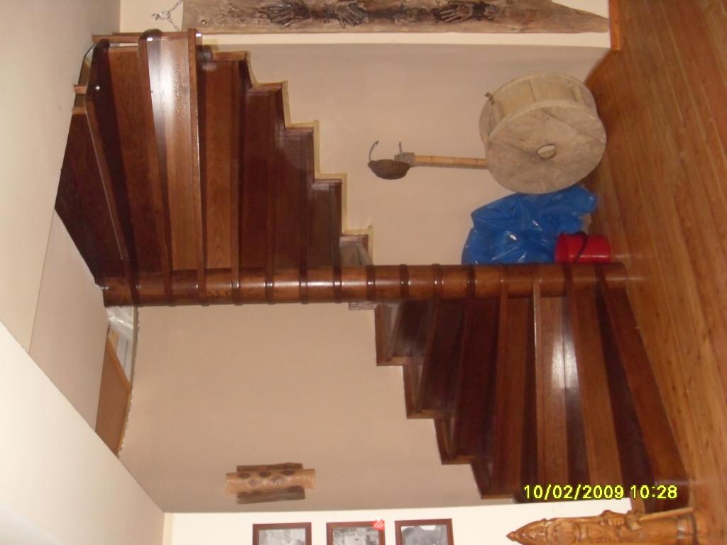 Kątowe ujęcie drewnianych schodów z ciemnego drewna z widoczną balustradą, na górze schodów leżą przedmioty zakryte niebieską folią, obok znajduje się drewniana ściana.