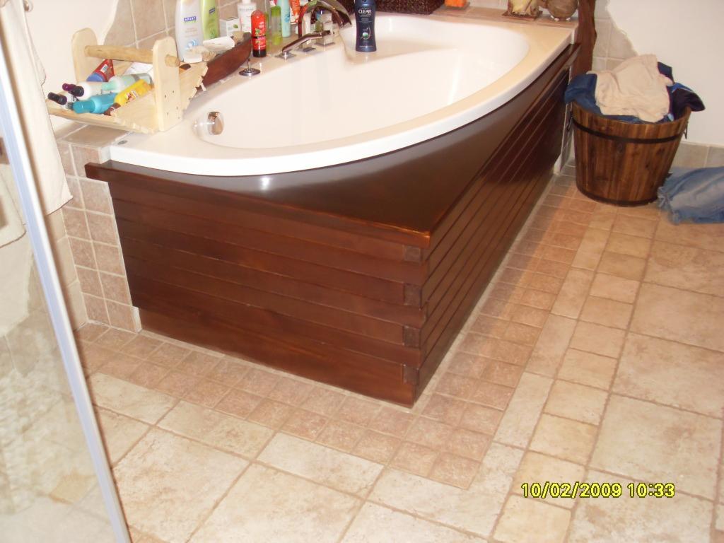 Obudowa wanny z ciemnego drewna, ułożona z poziomych desek, z widocznymi narożnikami, na tle beżowych płytek podłogowych i akcesoriów łazienkowych.