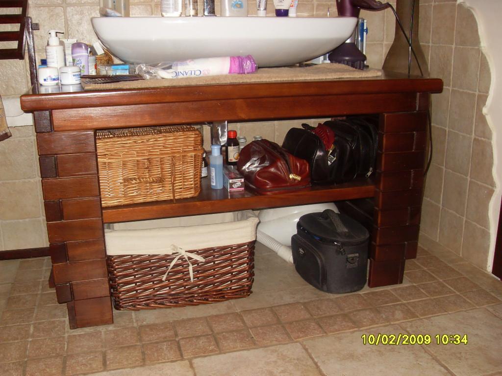 Ręcznie robiona, ciemnobrązowa szafka łazienkowa pod umywalką, wykończona elementami imitującymi cegły, z wiklinowymi koszami i torbami na półkach, na tle beżowych płytek.