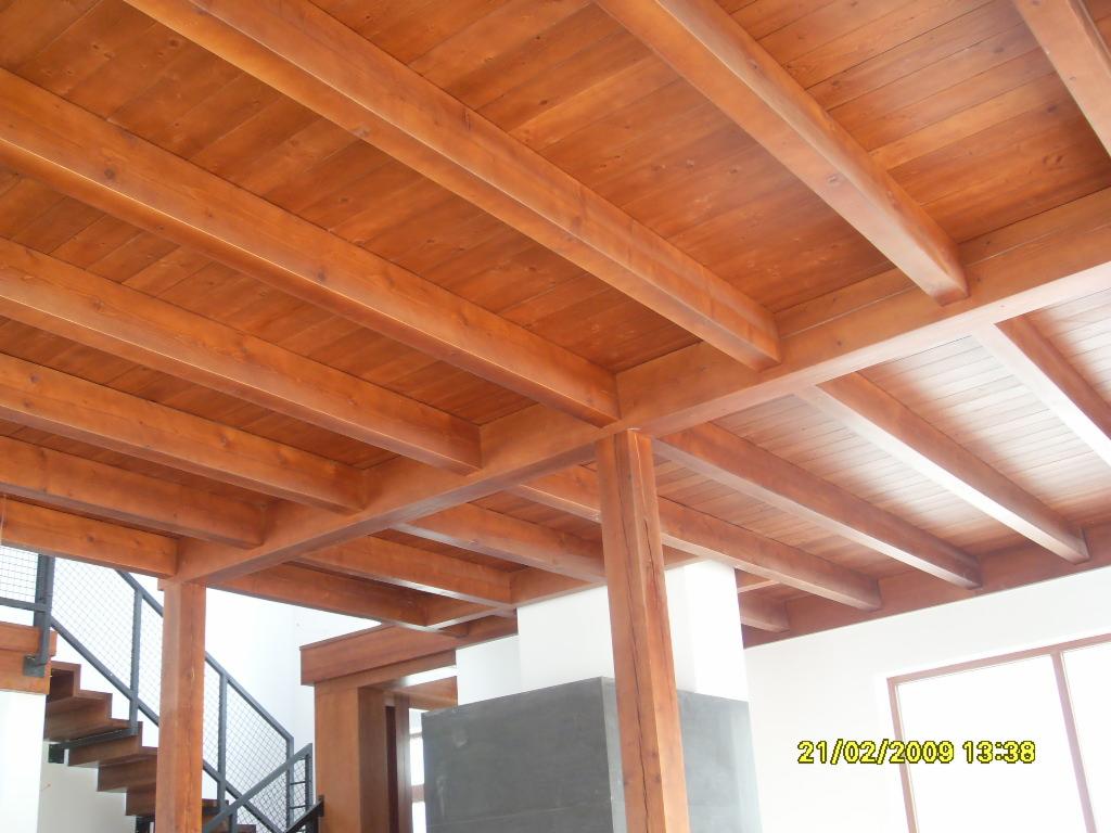 Drewniany sufit z belek stropowych w ciepłym odcieniu, widoczne schody z metalową balustradą w tle, fragment okna w prawym dolnym rogu.