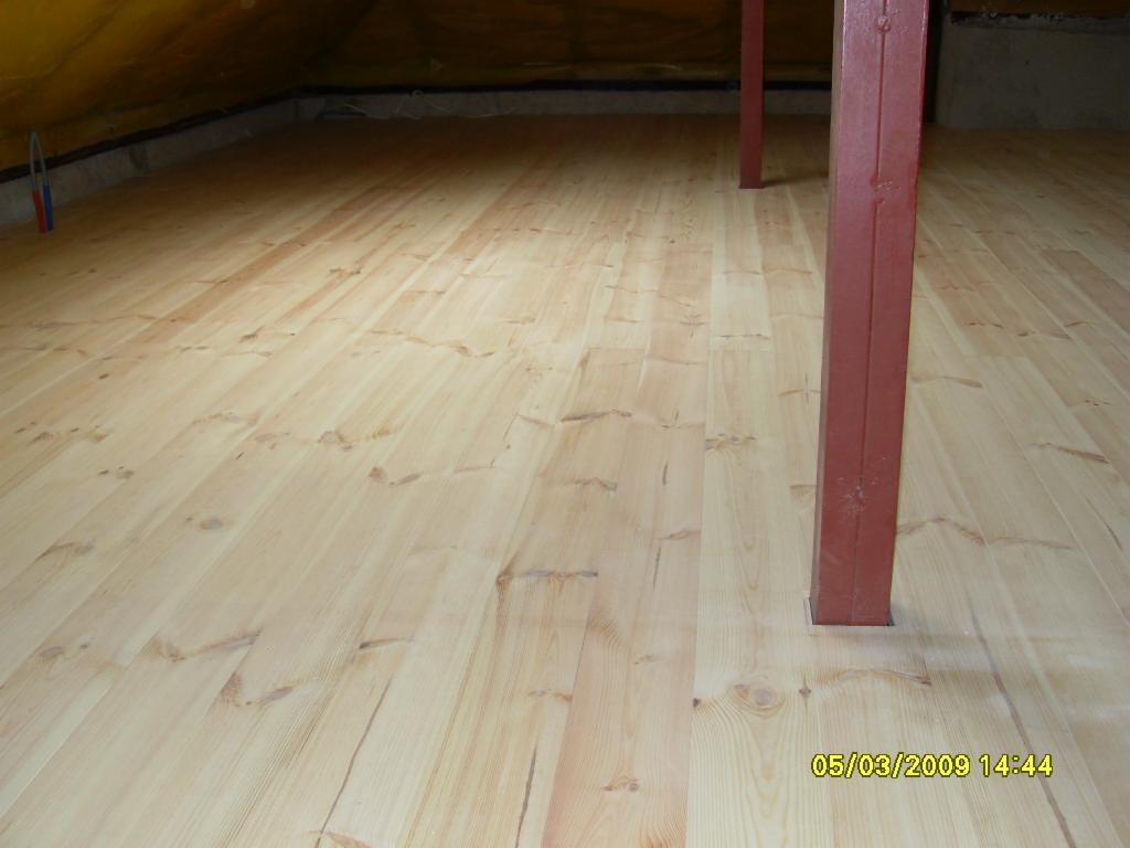 Podłoga z jasnych desek sosnowych w nowym poddaszu, widoczne czerwone słupy wspierające konstrukcję dachu, surowy stan wykończenia.