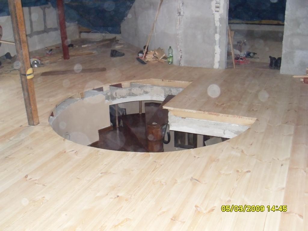 Montaż drewnianej podłogi z okrągłym otworem na schody w surowym wnętrzu poddasza, widoczne narzędzia i materiały budowlane.