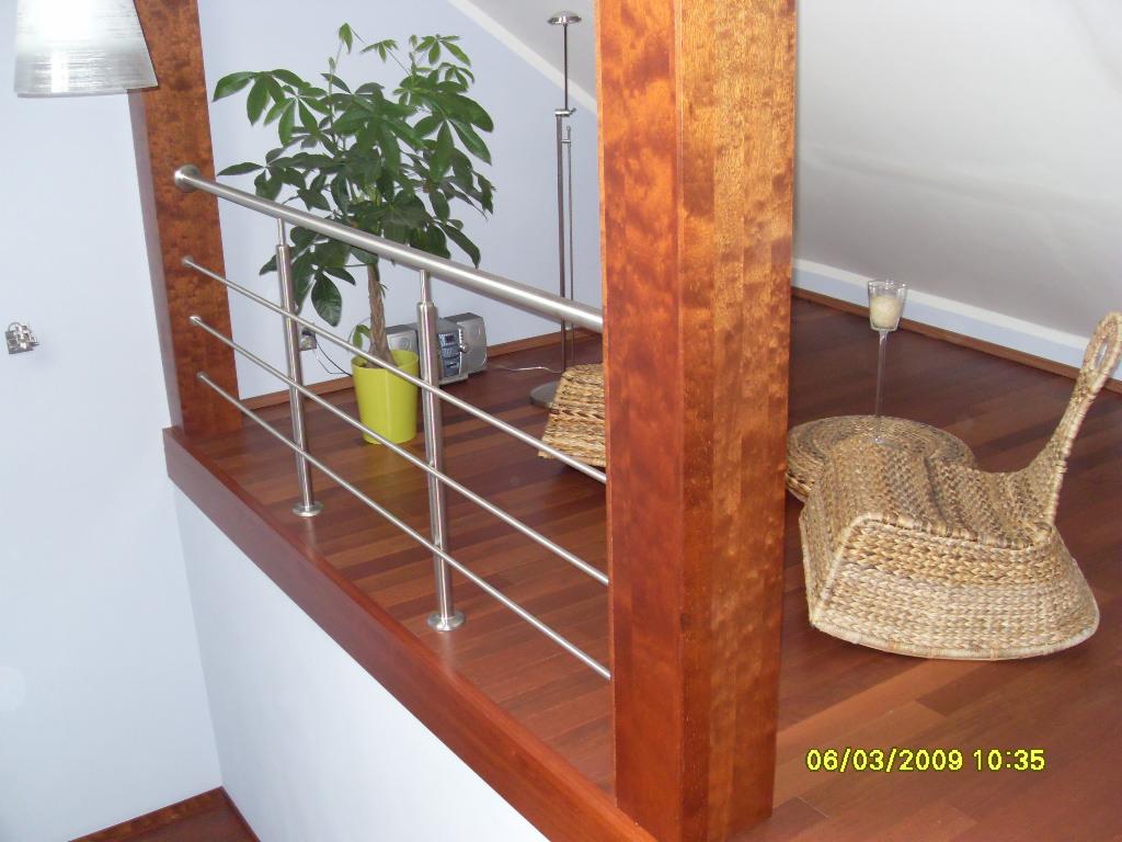 Drewniana podłoga z widocznymi schodami, balustradą z metalowymi elementami i drewnianą kolumną, wiklinowy fotel i roślina doniczkowa w tle.