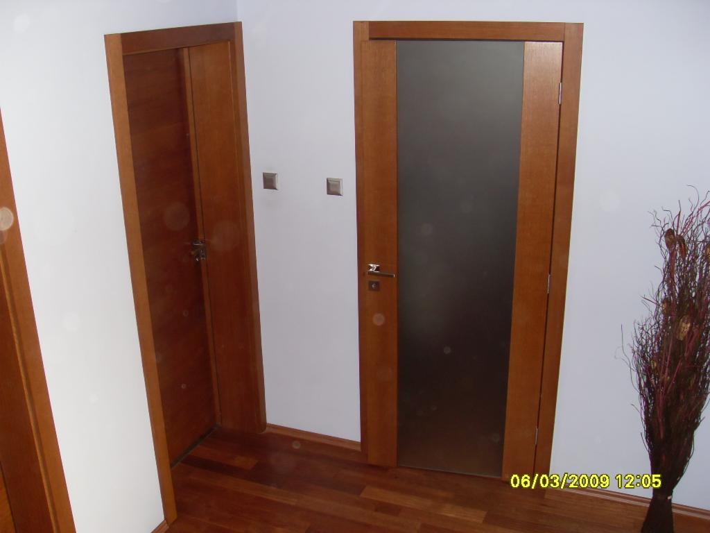 Dwa rodzaje drzwi wewnętrznych: pełne drewniane i z matową szybą, w brązowych ramach, na tle białych ścian i drewnianej podłogi, obok dekoracyjna sucha trawa w wazonie.