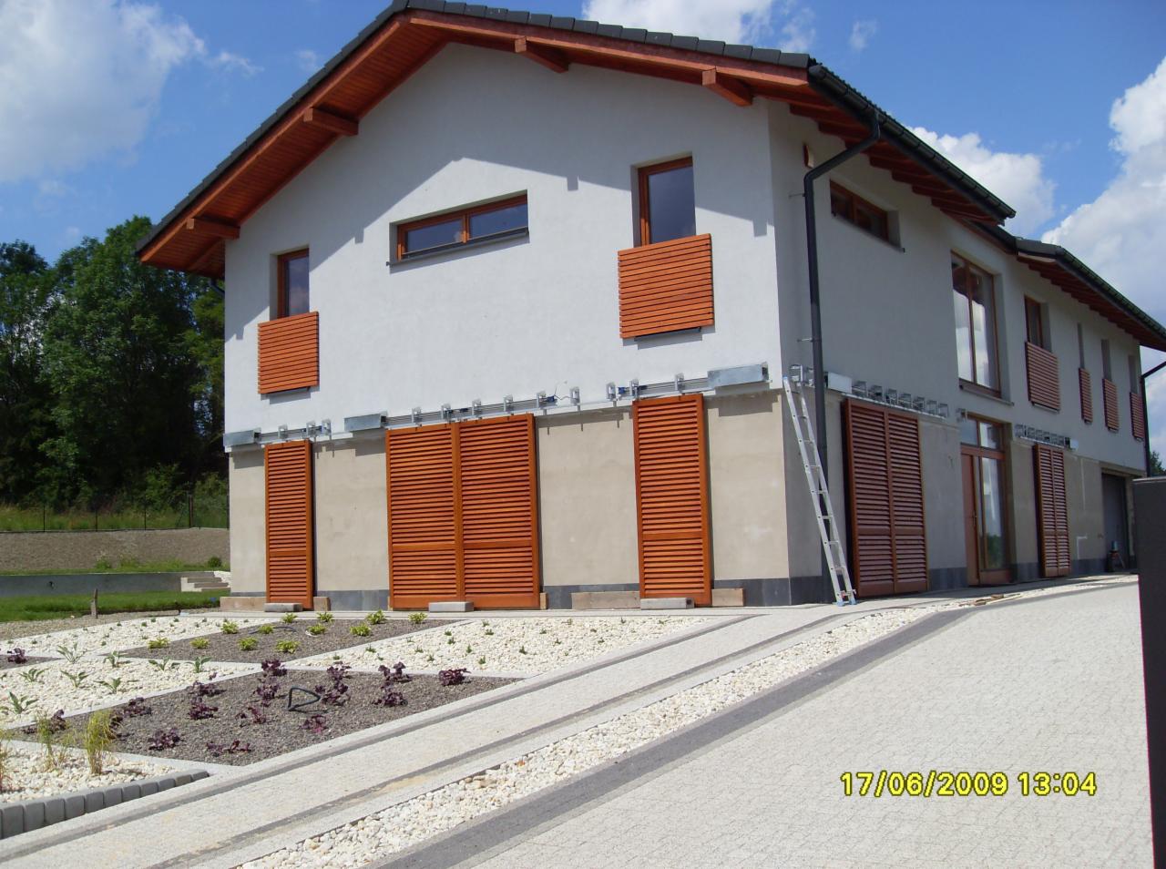 Nowoczesny dom z drewnianymi żaluzjami zewnętrznymi, zamontowanymi na szynach, częściowo zasłoniętymi, z aluminiową drabiną opartą o ścianę i zagospodarowanym ogrodem z widoczną datą wykonania...