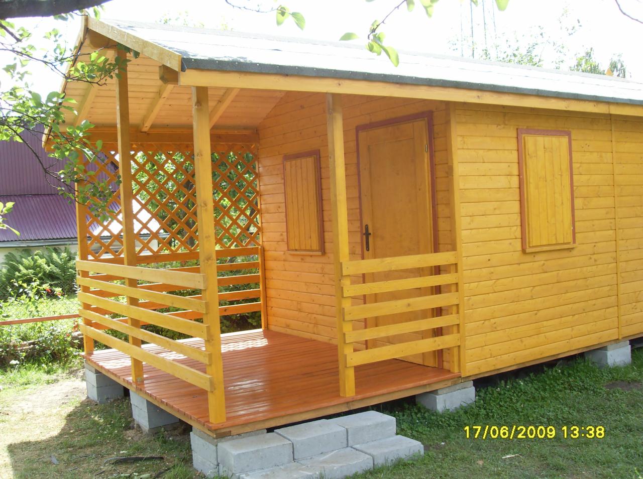 Żółty drewniany domek letniskowy z zadaszoną werandą, widoczne detale konstrukcji drewnianej i betonowe bloczki fundamentowe