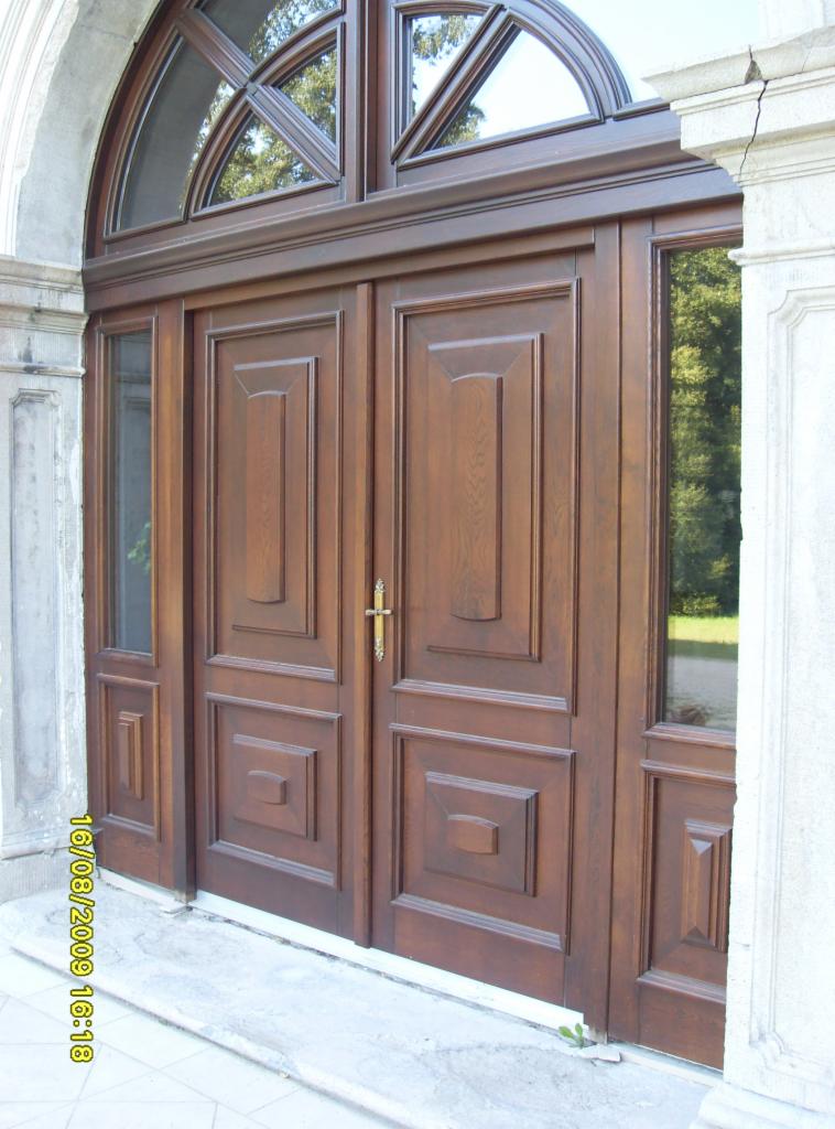 Dwuskrzydłowe, ciemnobrązowe drzwi wejściowe z drewna z dekoracyjnymi płycinami i przeszklonym nadświetlem łukowym, zamontowane w kamiennej fasadzie budynku.