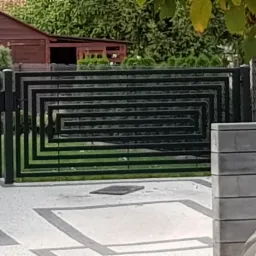 Nowoczesna brama posesyjna z czarnych, prostokątnych profili, tworząca geometryczny wzór labiryntu, na tle zielonego trawnika i fragmentu budynku gospodarczego.