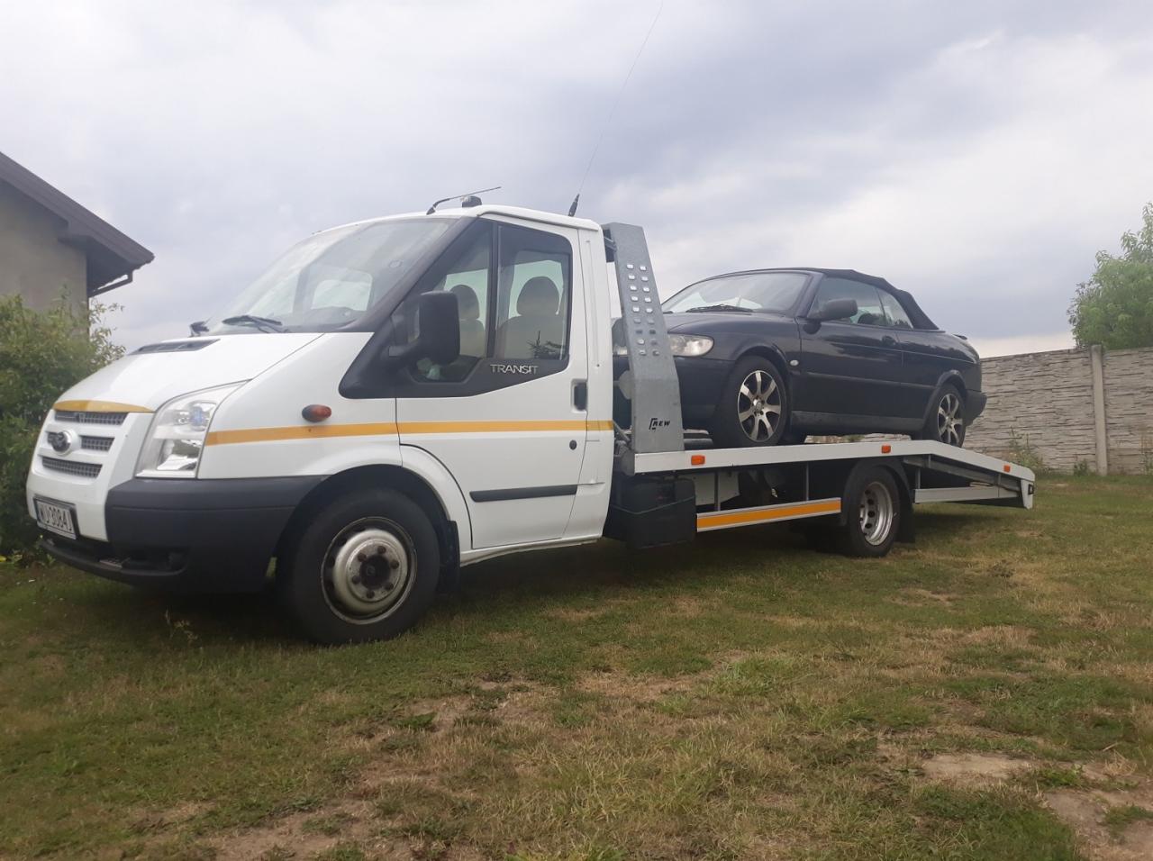 Biała laweta Ford Transit z czarnym Saabem cabrio na platformie, zaparkowana na trawie obok betonowego ogrodzenia i budynku. Pochmurne niebo.