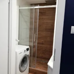 Kompaktowa łazienka z kabiną prysznicową ze szklanymi przesuwnymi drzwiami i drewnopodobną okładziną ścienną oraz pralką wciśniętą między ścianę a kabinę.