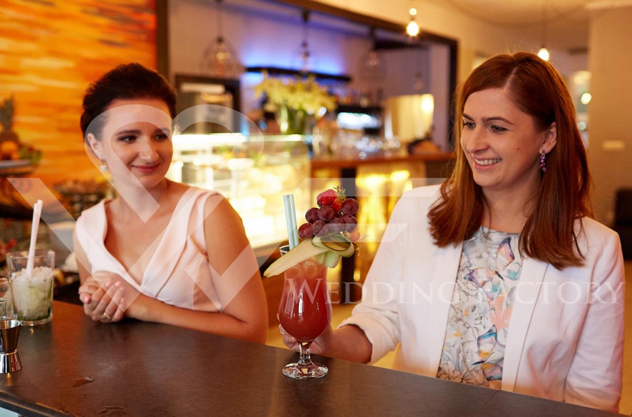 Dwie uśmiechnięte kobiety przy barze, jedna trzyma koktajl z owocową dekoracją, w tle ekspozycja z kwiatami i deserami.