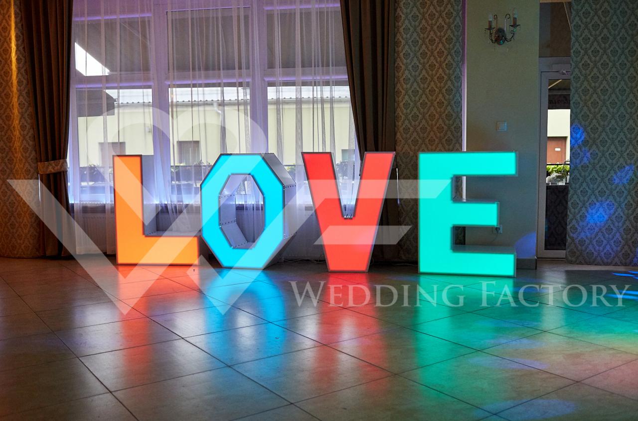 Podświetlane, kolorowe litery układające się w słowo LOVE, ustawione na podłodze w sali z oknem w tle. Widoczny napis WEDDING FACTORY.