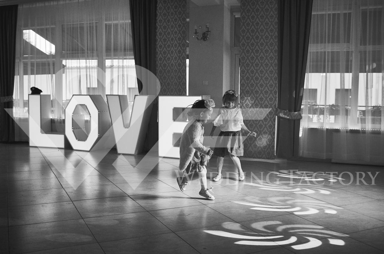 Dwie małe dziewczynki w sukienkach bawią się na parkiecie w sali z dekoracjami świetlnymi i dużym napisem LOVE w tle, czarno-białe zdjęcie.