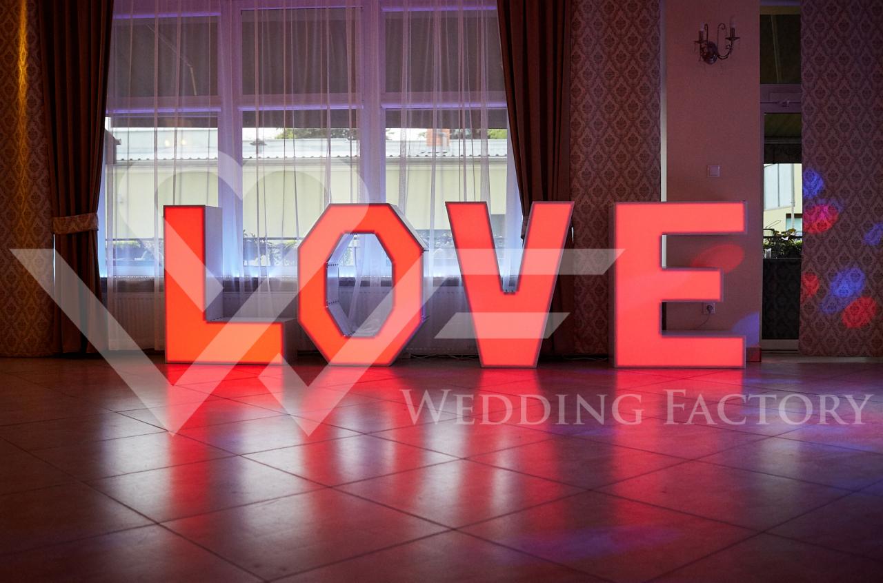 Podświetlane na czerwono litery układające się w słowo LOVE, ustawione na brązowej podłodze w sali z oknami i wzorzystą tapetą, z logo Wedding Factory na dole.