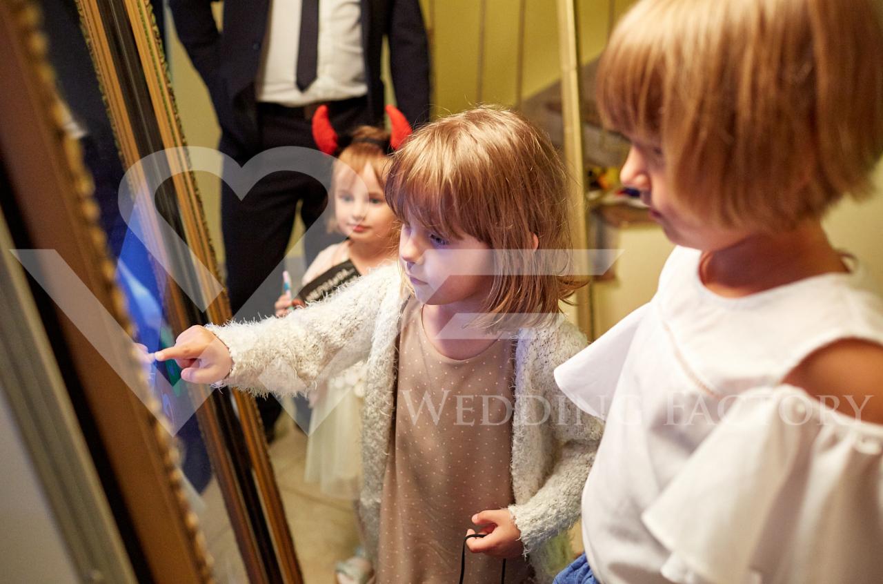 Dwie dziewczynki w jasnych ubraniach dotykają ekranu interaktywnej fotobudki, w tle widoczna postać mężczyzny w garniturze i inna dziewczynka z rogami diabła na głowie.