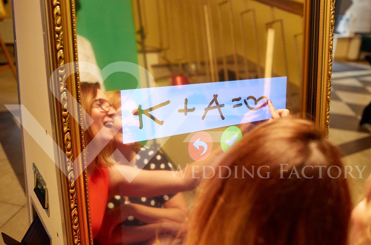 Osoby korzystające z interaktywnej fotobudki z ramą w stylu vintage, na ekranie dotykowym widnieje napis 'K+A=♥' oraz logo 'Wedding Factory'.