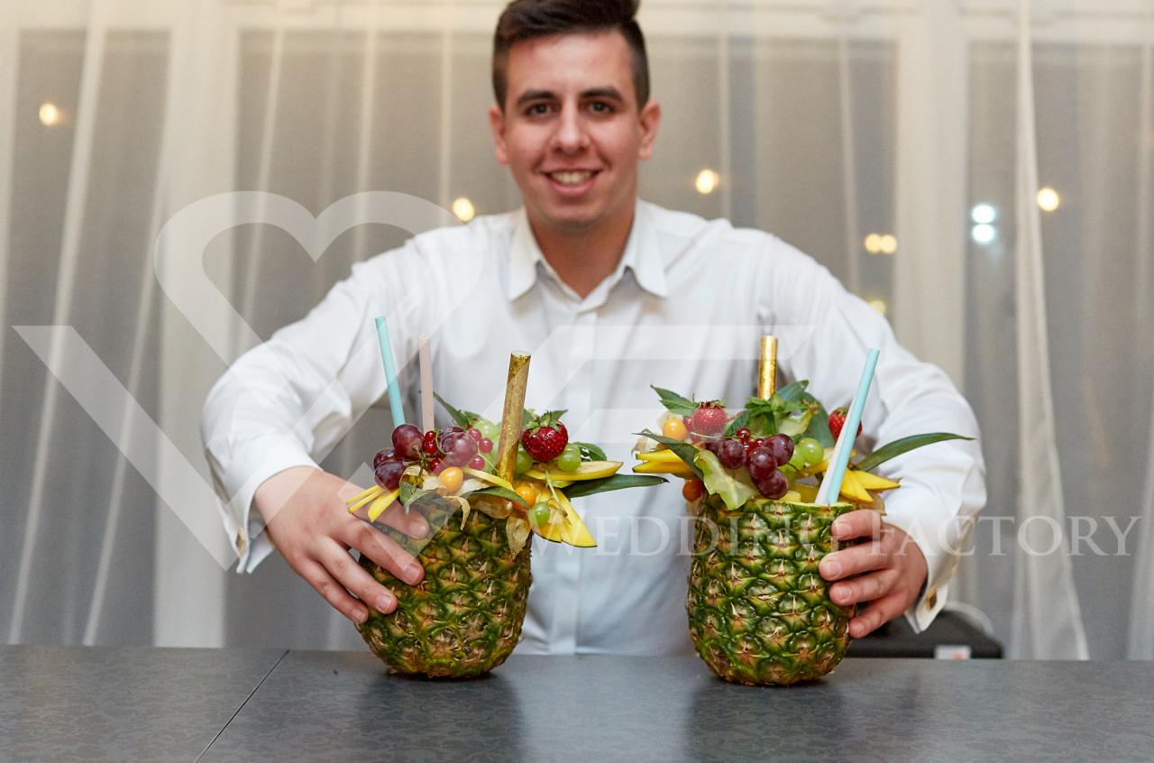Uśmiechnięty barman prezentuje dwa koktajle owocowe w wydrążonych ananasach, udekorowane winogronami, truskawkami i słomkami, na szarym blacie.