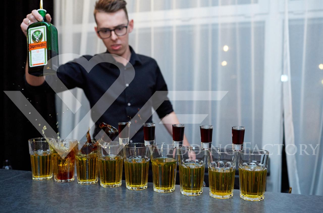 Barman w czarnej koszuli wlewa Jägermeistera do rzędu szklanek z żółtym płynem, tworząc efektowne rozbryzgi, z rzędem kieliszków w tle.