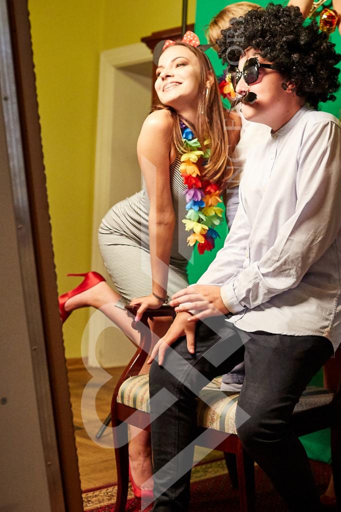 Para w przebraniach pozuje do zdjęcia w fotobudce: kobieta w sukience w paski z kwiatowym naszyjnikiem i opaską z uszami Myszki Miki, mężczyzna w peruce afro i okularach z wąsami.