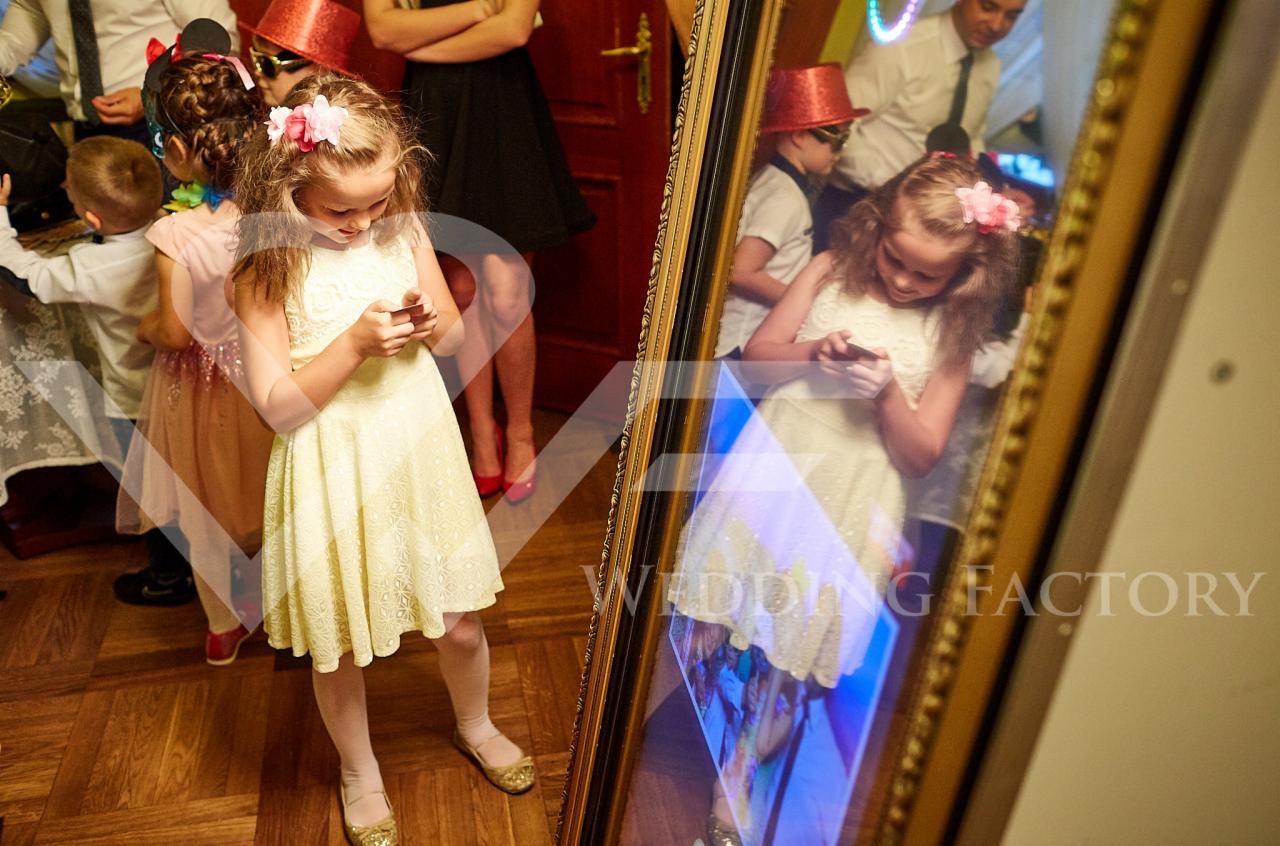Dziewczynka w żółtej sukience z kwiatem we włosach korzysta z fotobudki, trzymając telefon komórkowy, widoczna w lustrze fotobudki z innymi osobami w tle.