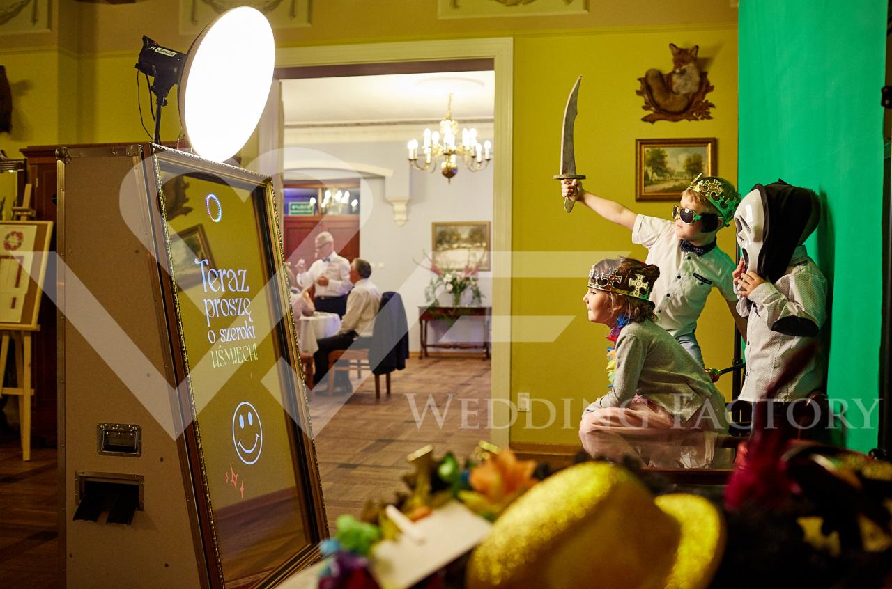 Trójka dzieci w przebraniach pozuje do zdjęcia w fotobudce z napisem 'Teraz proszę o szeroki uśmiech!' na tle eleganckiego wnętrza sali balowej z gośćmi weselnymi.