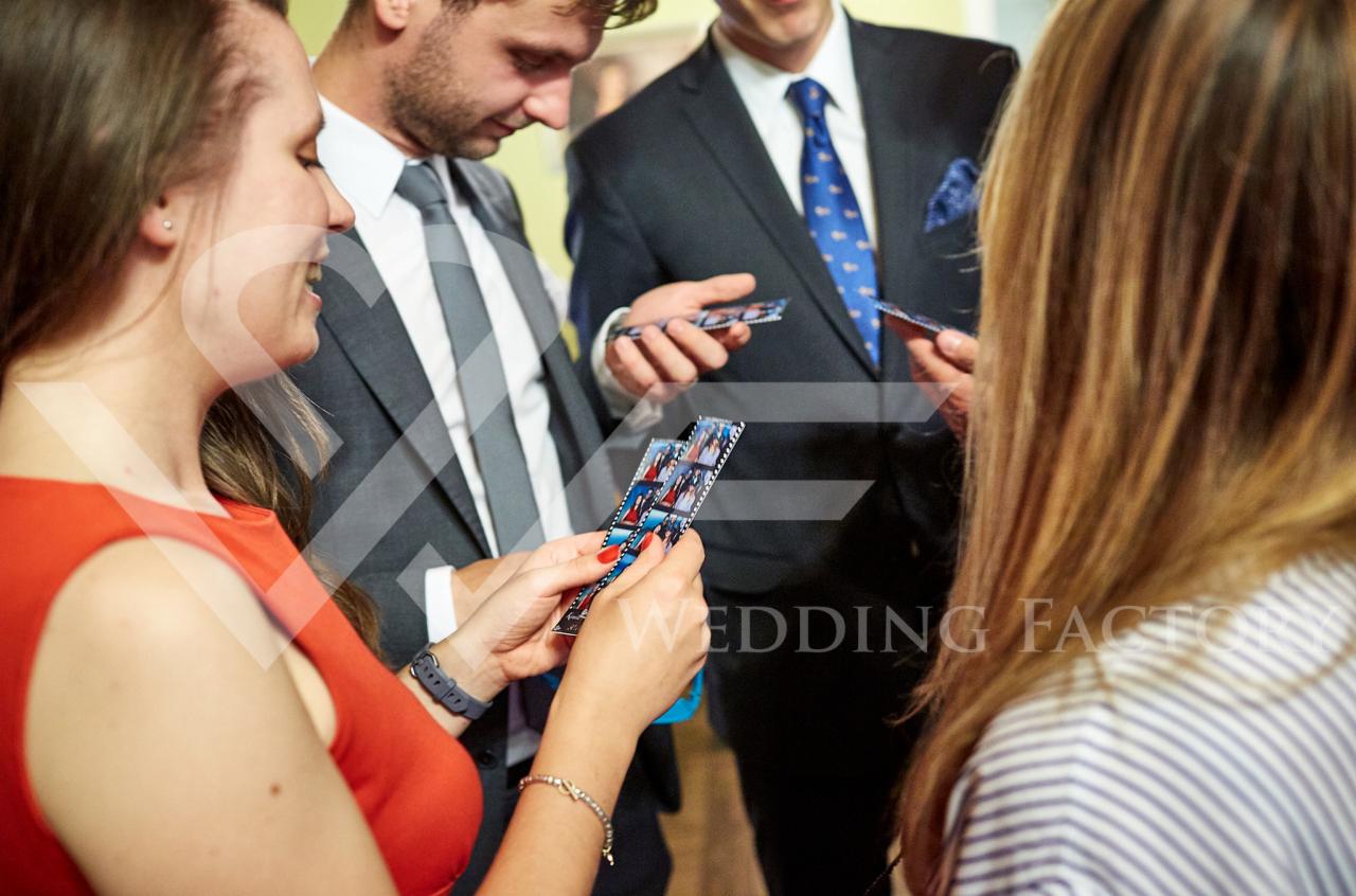 Grupa osób w Siedlcach ogląda paski ze zdjęciami z fotobudki, mężczyźni w garniturach, kobieta w czerwonej sukience, w tle inny mężczyzna w garniturze z niebieskim krawatem.