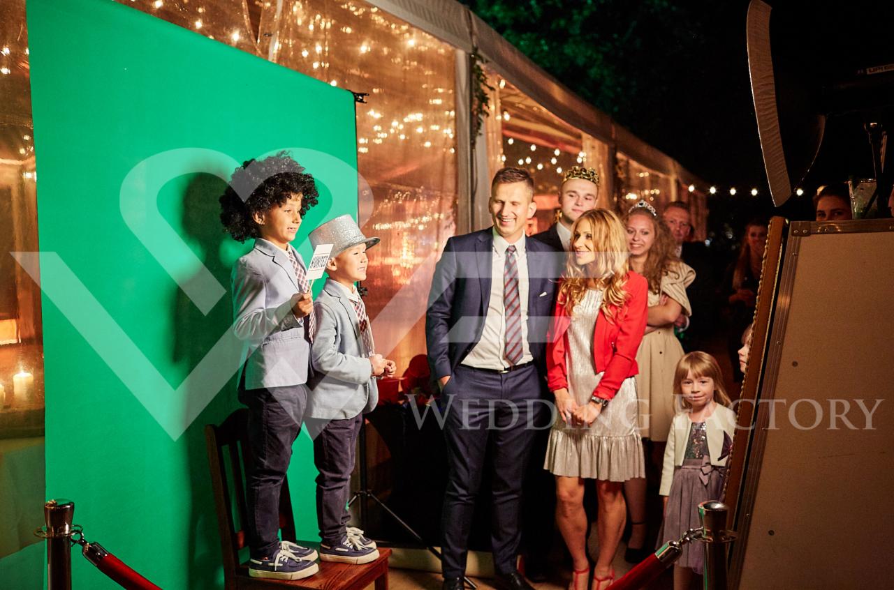 Goście weselni pozują do zdjęcia w fotobudce z zielonym tłem, chłopcy w przebraniach stoją na krześle, a para młoda i inni goście czekają na swoją kolej.